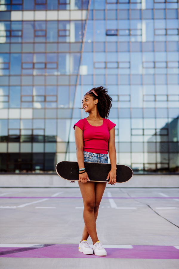 Multiracial teenage girl with backpack and skateboard, walking in city during a summer day.