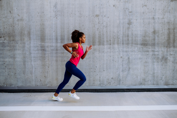 Young multiracial girl jogging in city, in front of a concrete wall, side view.