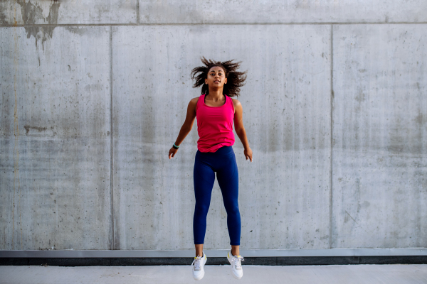 Young multiracial girl jogging and jumping in city, in front of a concrete wall, side view.