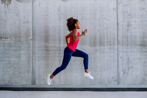 Young multiracial girl jogging and jumping in city, in front of a concrete wall, side view.