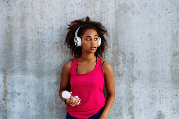 Young multiracial girl in sportswear resting after jogging in city, drinking water from a sustainable bottle and listening music in headphones.