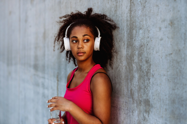 Young multiracial girl in sportswear resting after jogging in city, drinking water from a sustainable bottle and listening music in headphones.