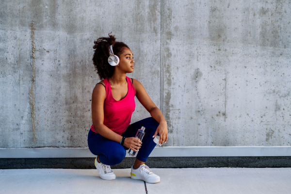 Young multiracial girl in sportswear resting after jogging in city, drinking water from a sustainable bottle and listening music in headphones.