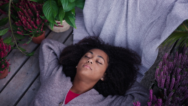 A young woman lying on patio with closed eyes having peaceful moment. Beautiful african american woman resting, in connection with plants around her.