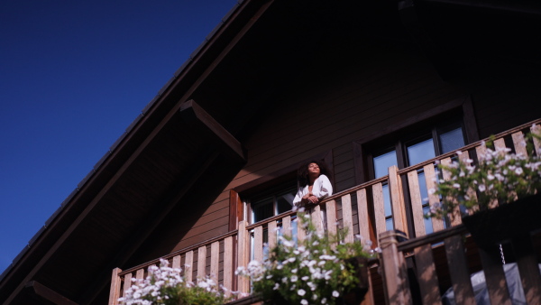 A peaceful morning for young woman standing on balcony in bathrobe, looking at beautiful nature around her, smiling.