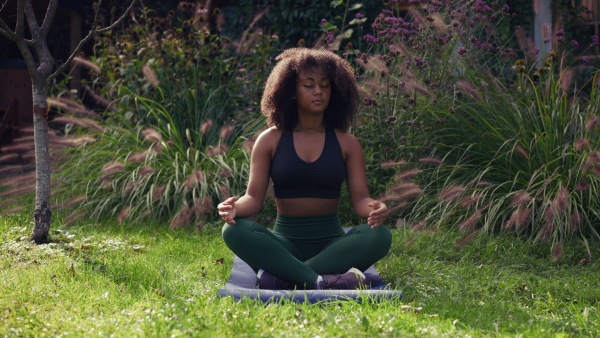 A young woman meditating outdoors, sitting on lawn in the garden with closed eyes in yoga pose. Beautiful african american woman connecting with herself and nature.