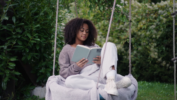 A young woman sitting on swing with book, in the middle of lush foliage. patio with cup of tea, having peaceful moment. Beautiful african american woman reading in the middle of nature.