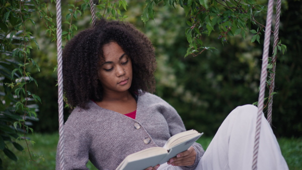 A young woman sitting on swing with book, in the middle of lush foliage. patio with cup of tea, having peaceful moment. Beautiful african american woman reading in the middle of nature.