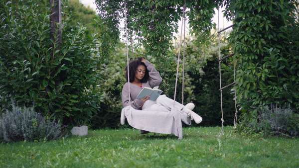 A young woman sitting on swing with book, in the middle of lush foliage. patio with cup of tea, having peaceful moment. Beautiful african american woman reading in the middle of nature.