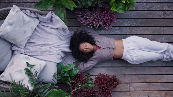 A young woman lying on patio with closed eyes having peaceful moment. Beautiful african american woman resting, in connection with plants around her.