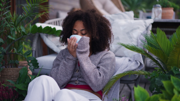 A young woman sitting on patio with cup of tea, having peaceful moment. Beautiful african american woman resting, looking at nature around her.