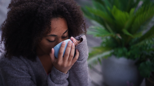 A young woman sitting on patio with cup of tea, having peaceful moment. Beautiful african american woman resting, looking at nature around her.