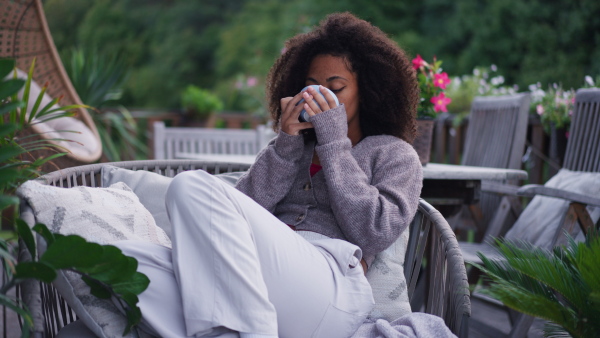 A young woman sitting on patio with cup of tea, having peaceful moment. Beautiful african american woman resting, looking at nature around her.