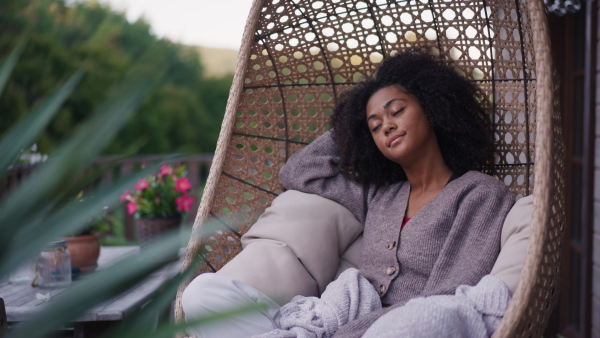 A young woman sitting outdoors in hanging chair on patio, having peaceful moment. Beautiful african american woman resting with closed eyes, snuggling under blanket.