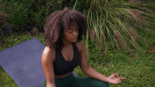 A young woman meditating outdoors, sitting on lawn in the garden with closed eyes in yoga pose. Beautiful african american woman connecting with herself and nature.