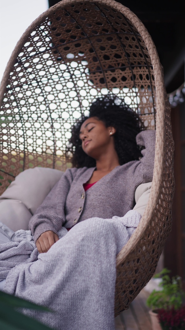 A young woman sitting outdoors in hanging chair on patio, having peaceful moment. Beautiful african american woman resting with closed eyes, snuggling under blanket.