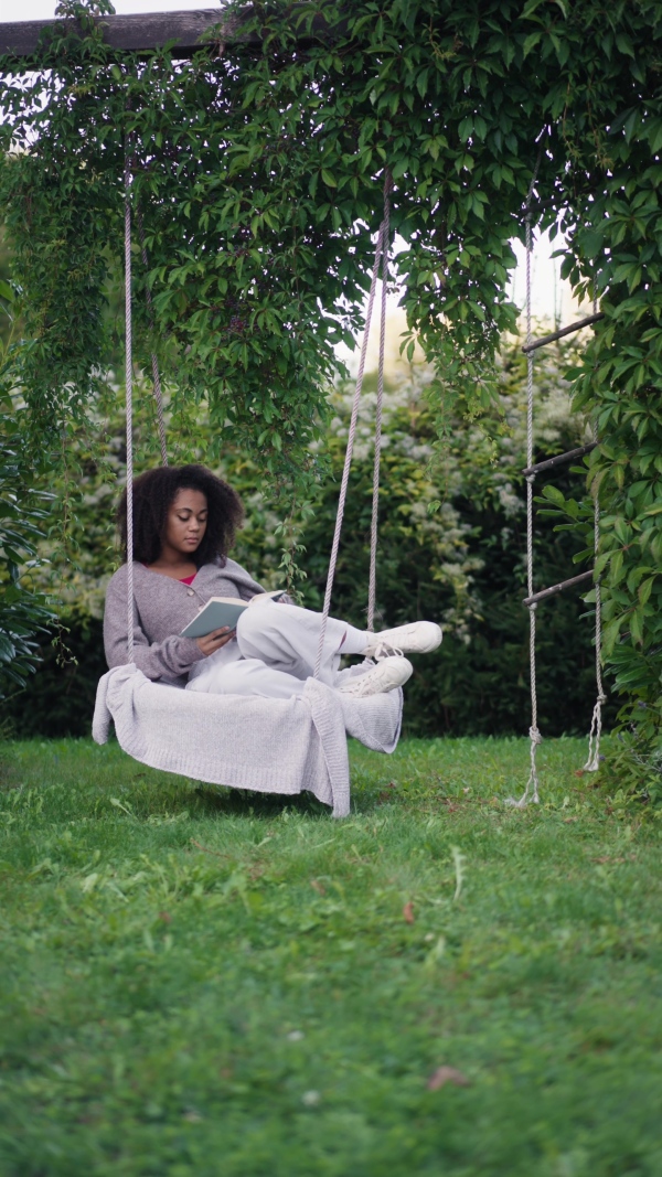 A young woman sitting on swing with book, in the middle of lush foliage. patio with cup of tea, having peaceful moment. Beautiful african american woman reading in the middle of nature.