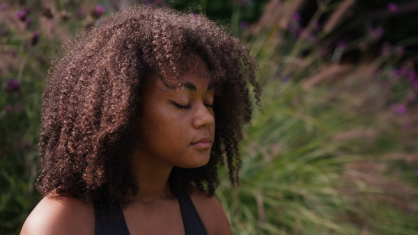 A young woman meditating outdoors, sitting on lawn in the garden with closed eyes in yoga pose. Beautiful african american woman connecting with herself and nature.