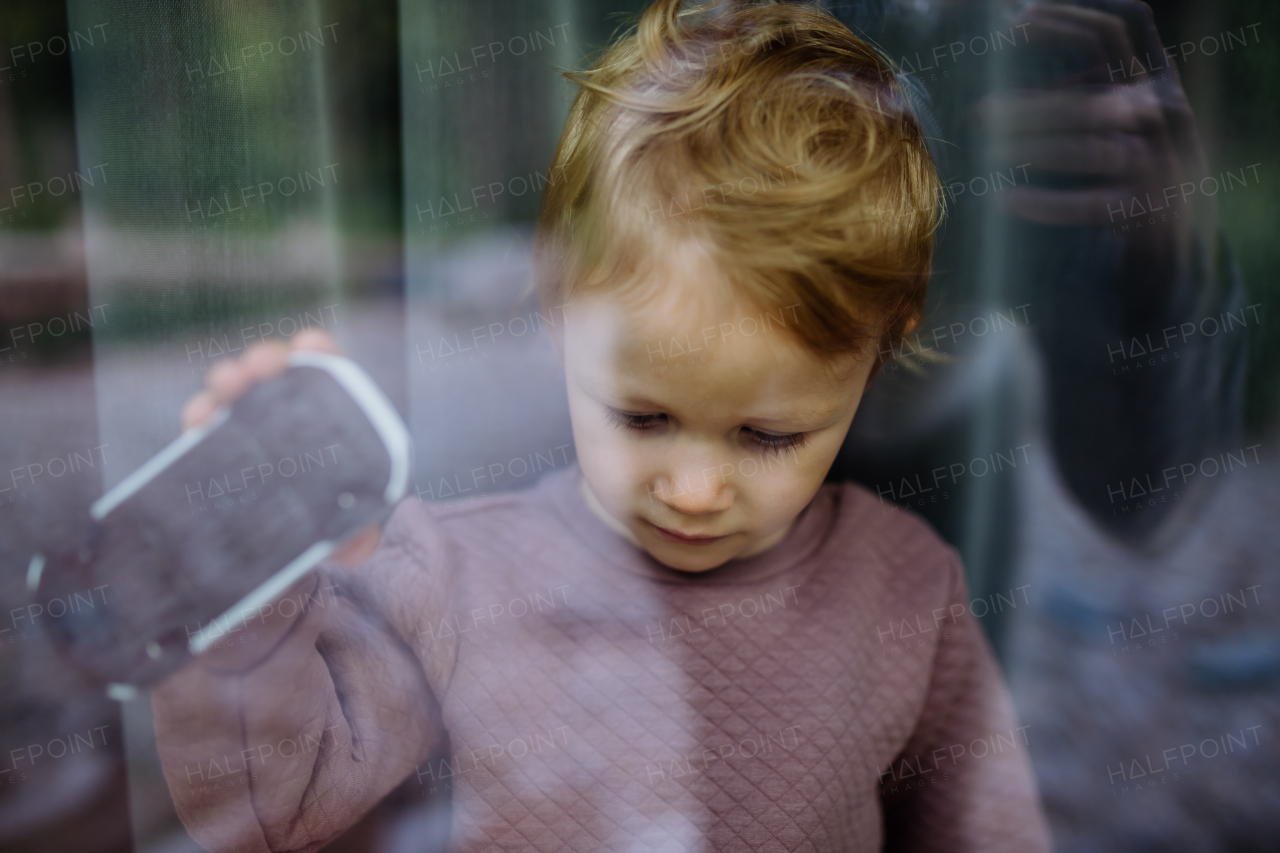 Sad little child standing alone with car toy behind the window, photo trough glass.