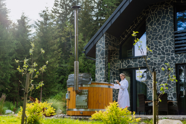 Young woman in bathrobe, checking temperature with thermometer, ready for home spa procedure in hot tub outdoors. Wellness, body care, hygiene concept.