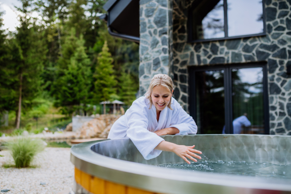 Young woman in bathrobe, checking temperature with thermometer, ready for home spa procedure in hot tub outdoors. Wellness, body care, hygiene concept.