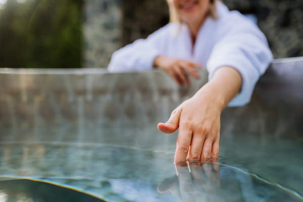 An unrecognizable woman in towel touching water, checking temperature, ready for home spa procedure in hot tub outdoors. Wellness, body care, hygiene concept.