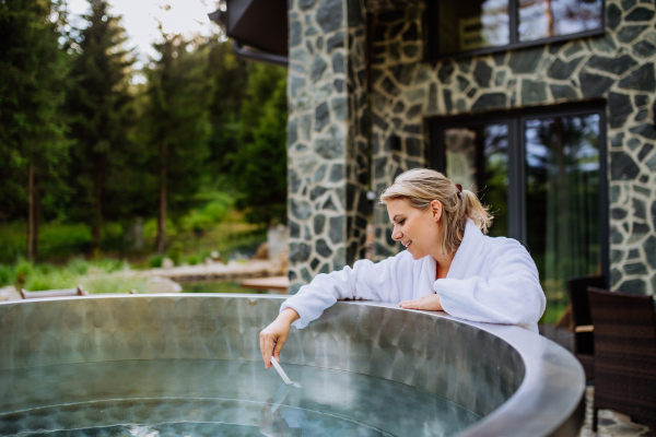 Young woman in bathrobe, checking temperature with thermometer, ready for home spa procedure in hot tub outdoors. Wellness, body care, hygiene concept.