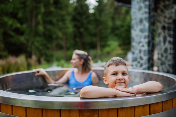 Mother with her little son enjoying bathing in a wooden barrel hot tub in the terrace of the cottage. Wooden bathtub with a fireplace to burn wood and heat water.