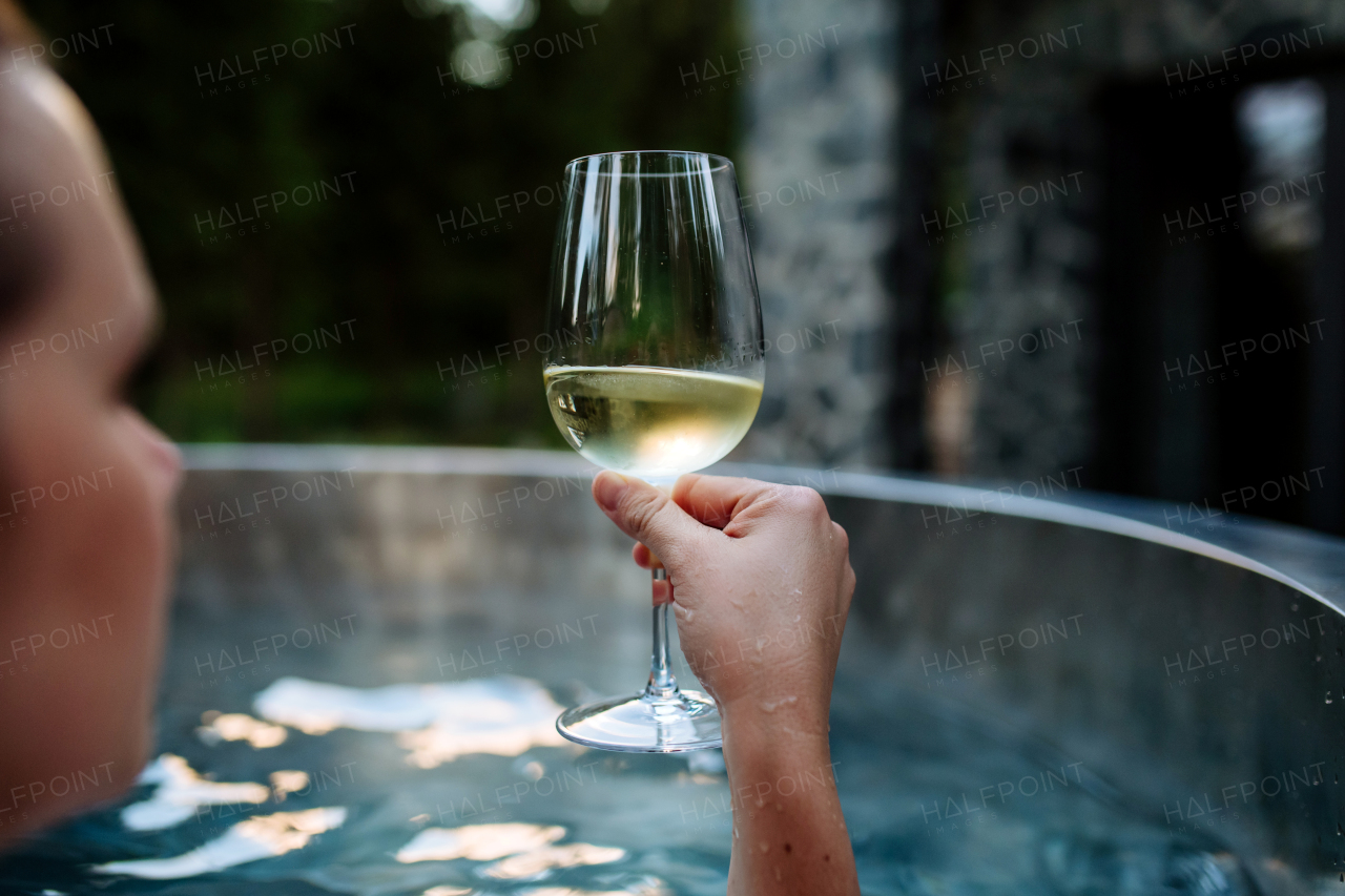 An unrecognizable young woman relaxing with glass of wine in hot tub outdoor in nature.