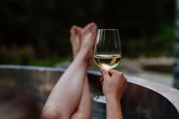 An unrecognizable young woman with feet up relaxing with glass of wine in hot tub outdoor in nature.