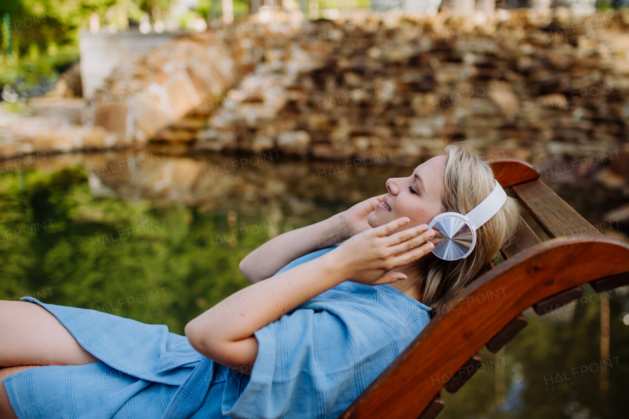 A relaxed woman wearing headphones listening to music sitting on a pier by natureal lake in summer