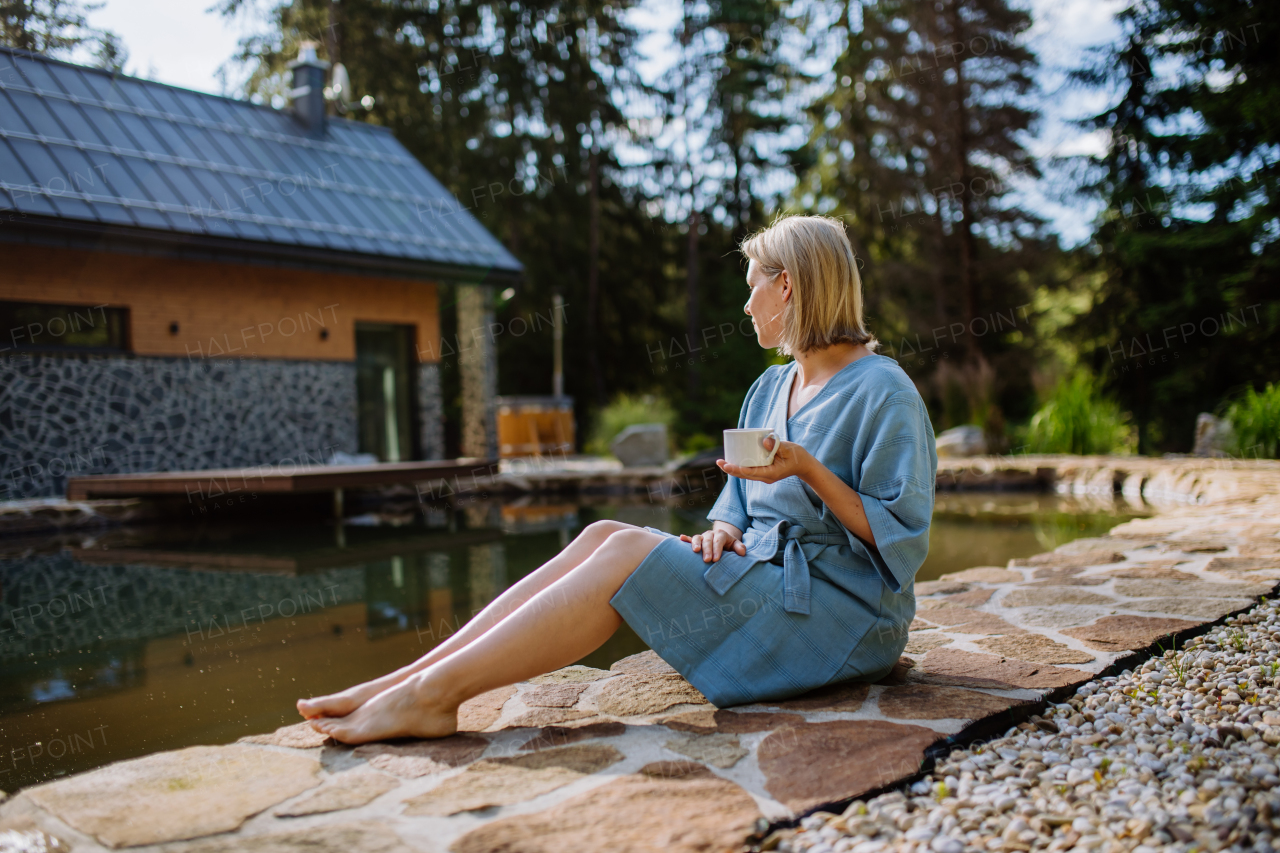 A happy young woman sitting by pond near cottege and enjoying cup of morning coffee on summer vacation in mountains. High angle view.