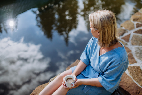 A happy young woman sitting by pond near cottege and enjoying cup of morning coffee on summer vacation in mountains.