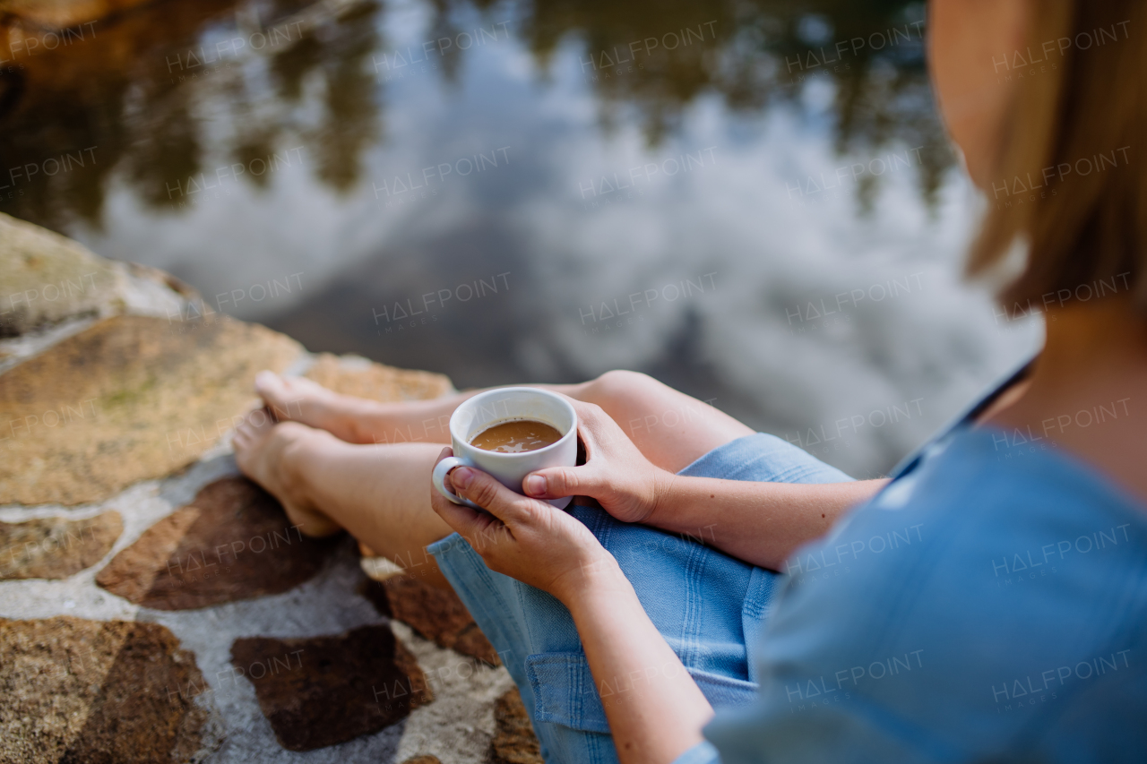 A happy young woman sitting by pond near cottege and enjoying cup of morning coffee on summer vacation in mountains. High angle view.