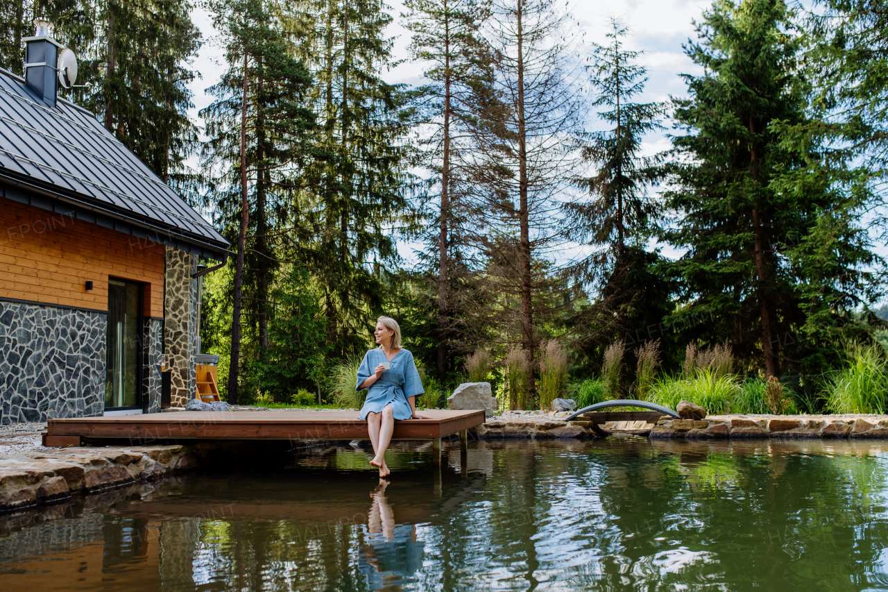 Woman, sitting on a pier,near private lake, relaxing, enjoying a cup of morning coffee on summer vacation in mountains.