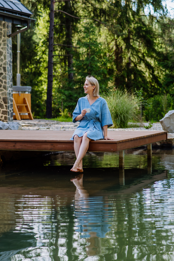 A happy young woman sitting by pond near cottege and enjoying cup of morning coffee on summer vacation in mountains. High angle view.