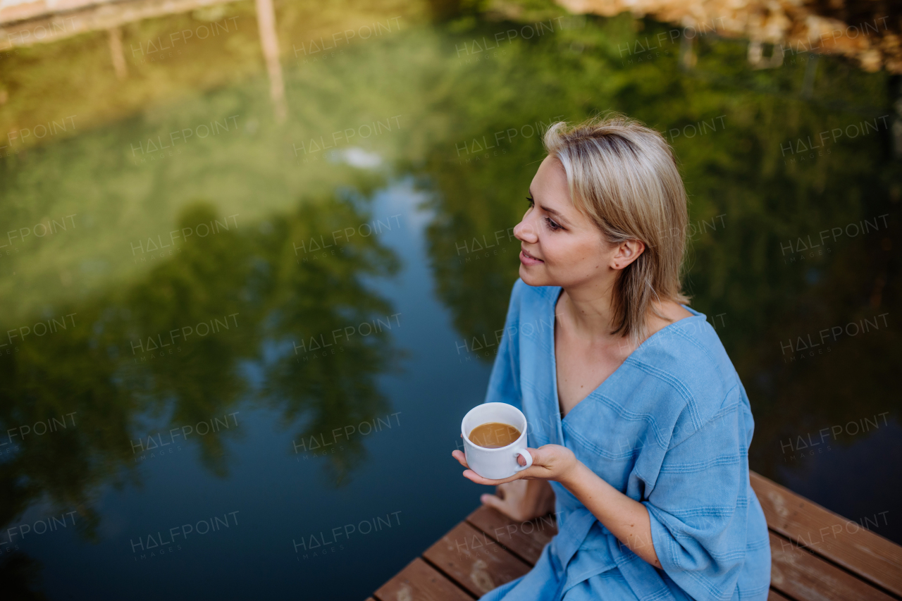 A happy young woman sitting by pond near cottege and enjoying cup of morning coffee on summer vacation in mountains. High angle view.