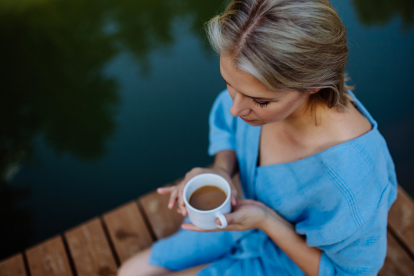 A happy young woman sitting by pond near cottege and enjoying cup of morning coffee on summer vacation in mountains. High angle view.