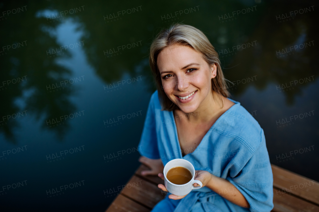 A happy young woman sitting by pond near cottege and enjoying cup of morning coffee on summer vacation in mountains. High angle view.