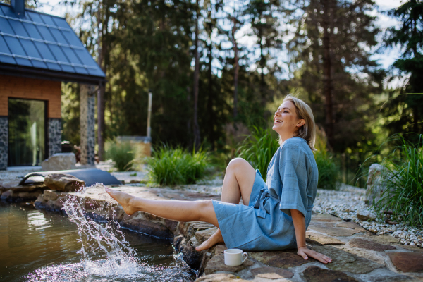 A happy young woman dipping her foot and splashing in pond near cottege, during hot sunny day on summer vacation in mountains.