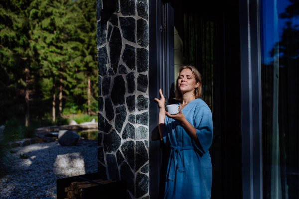 A happy young woman standing in garden near cottege and enjoying cup of morning coffee on summer vacation in mountains.