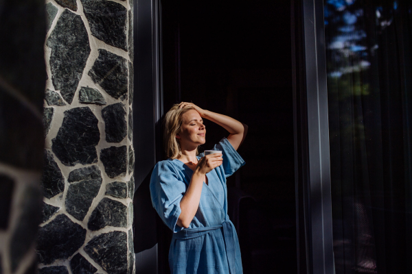 A happy young woman standing in garden near cottege and enjoying cup of morning coffee on summer vacation in mountains.