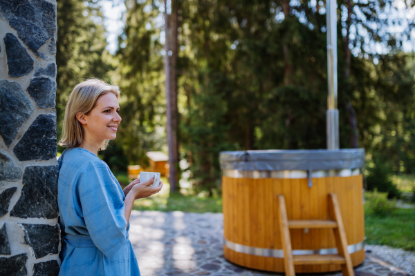 A happy young woman standing in garden near hot tub and enjoying cup of morning coffee on summer vacation in mountains.