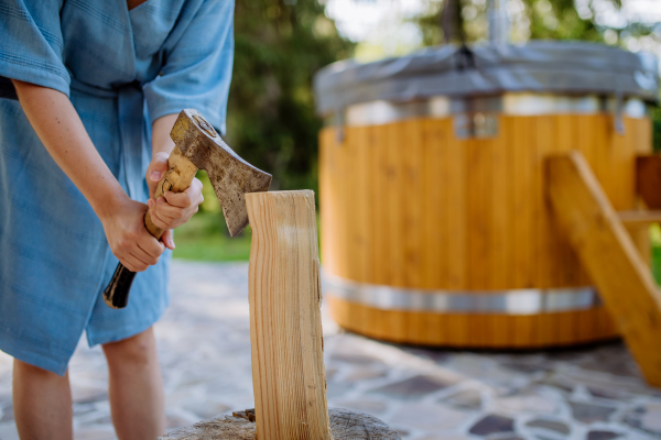 An unrecognizable woman chopping wood preparing it for for wooden bathtub with a fireplace to burn wood and heat water in backyard in mountains.