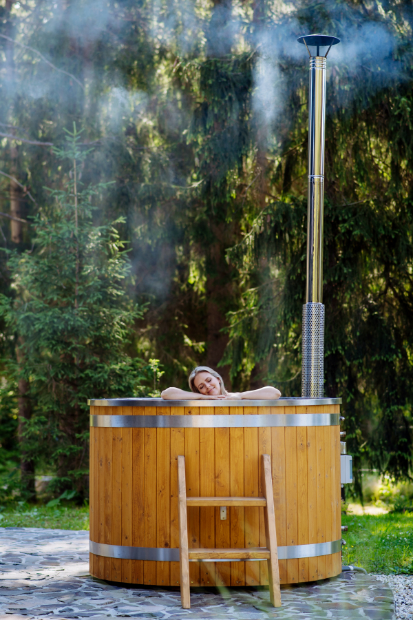 Young woman enjoying wooden bathtub with fireplace to burn wood and heat water in backyard in mountains.