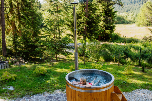 Young woman enjoying wooden bathtub with fireplace to burn wood and heat water in backyard in mountains.