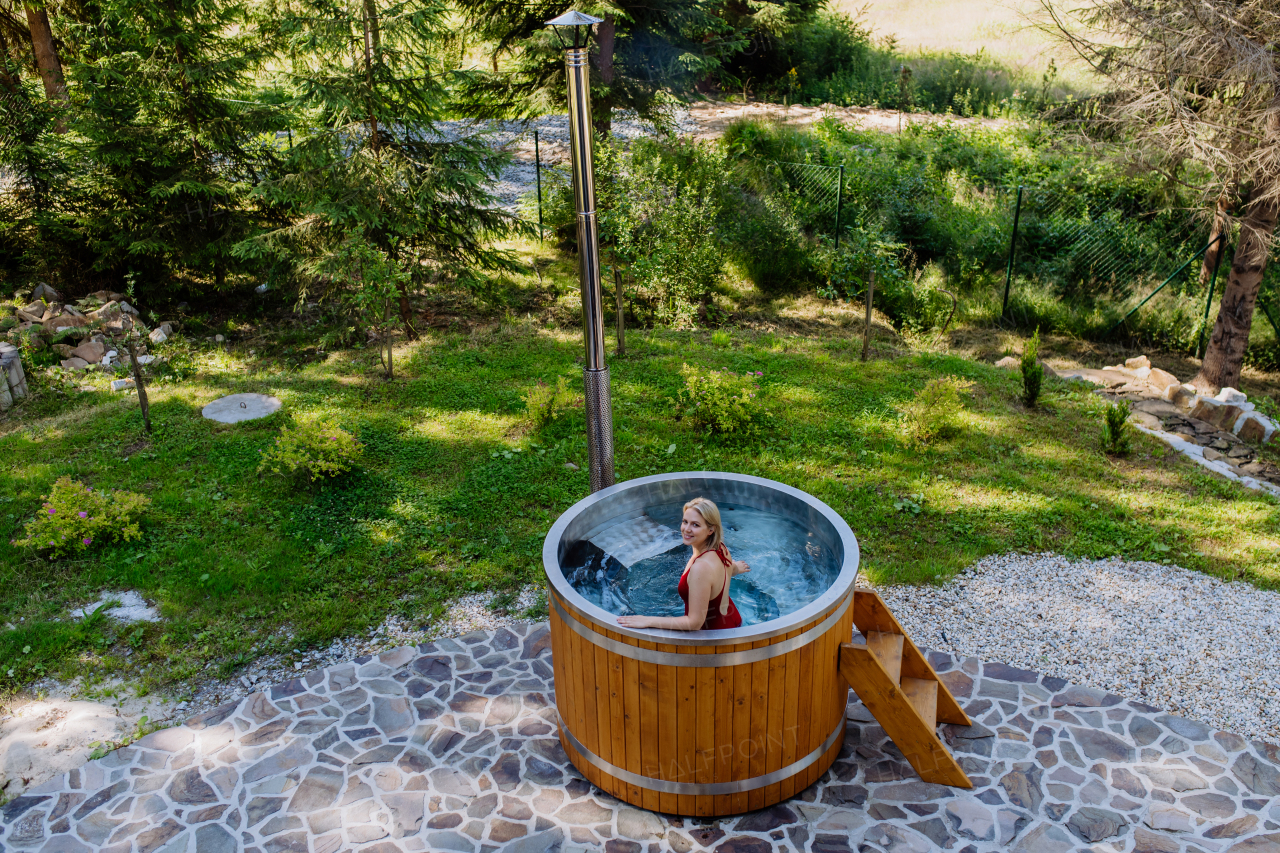 Young woman enjoying wooden bathtub with fireplace to burn wood and heat water in backyard in mountains. High angle view.