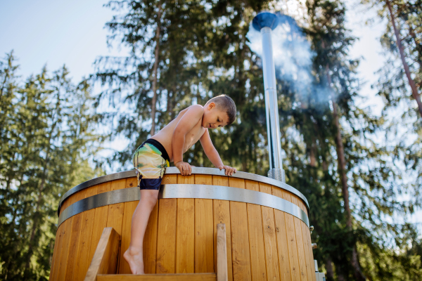 Little boy in swimsuit climbing in the outdoor wooden hot tub,surrounded by forest, during a sunny summer day. Low angle view.