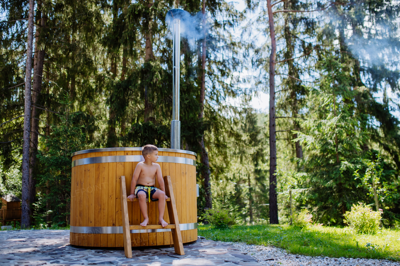 Little boy in swimsuit sitting and resting in outdoor wooden hot tub stairs,surrounded by forest, during sunny summer day.