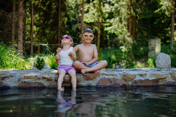 A cute girl and boy sitting together on the footpath by the lake with sun glasses, dangle their feet in the water, summer vacation concept.